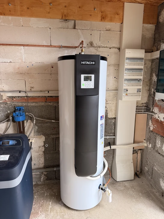 Un ballon thermodynamique blanc ou gris clair installé dans un local technique résidentiel, visible dans son contexte d'installation avec tuyauteries de raccordement, éventuellement accompagné d'indicateurs visuels subtils montrant la circulation thermique (vapeur ou condensation légère), illustrant une solution moderne et compacte de production d'eau chaude sanitaire.